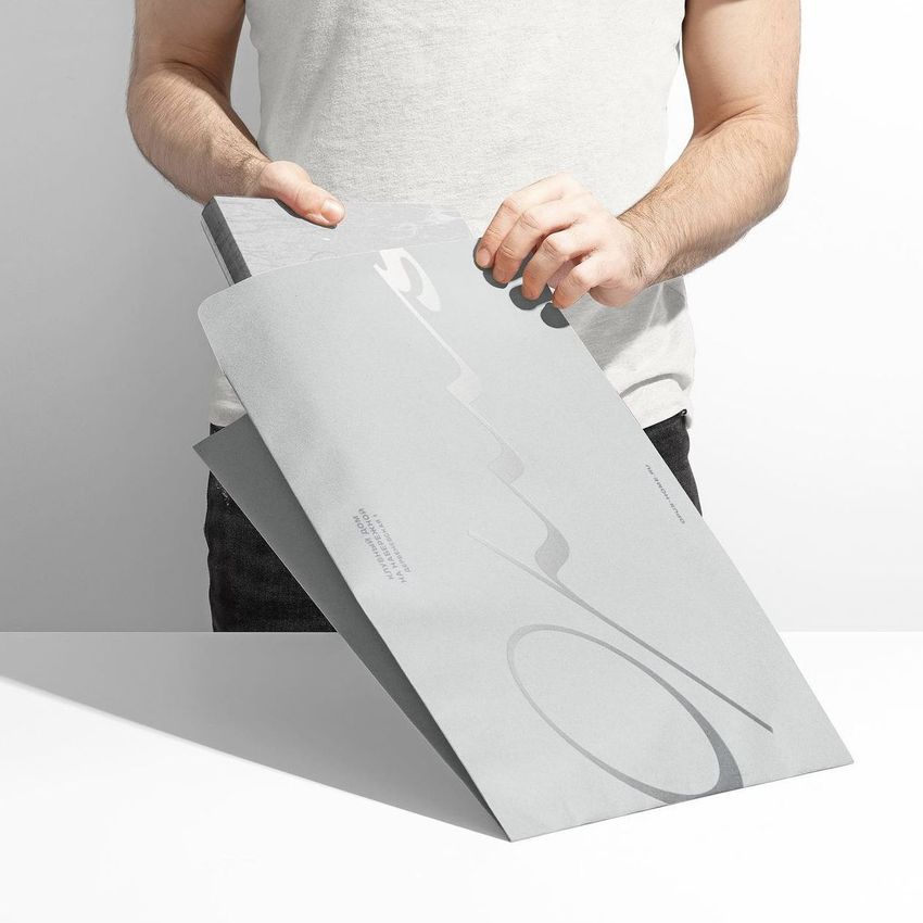 Person holding and opening a large gray kraft mailer mockup with minimalist abstract graphics, set against a plain white background.