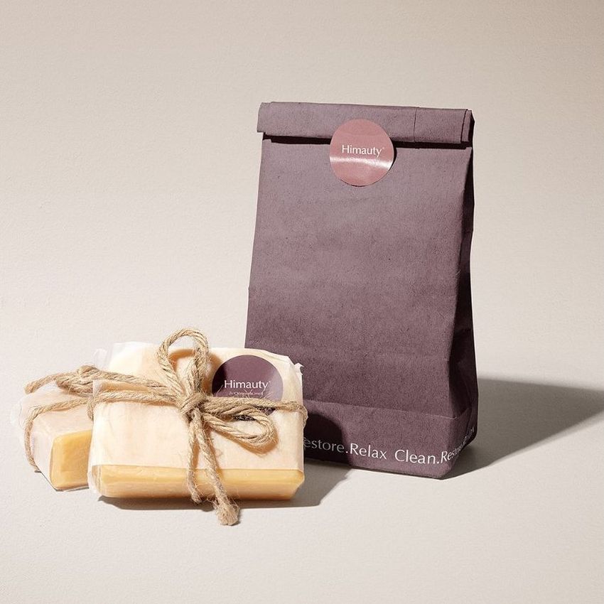 Handmade soap packaging mockup featuring a dark paper bag with a round label and two tied soap bars on a neutral surface.