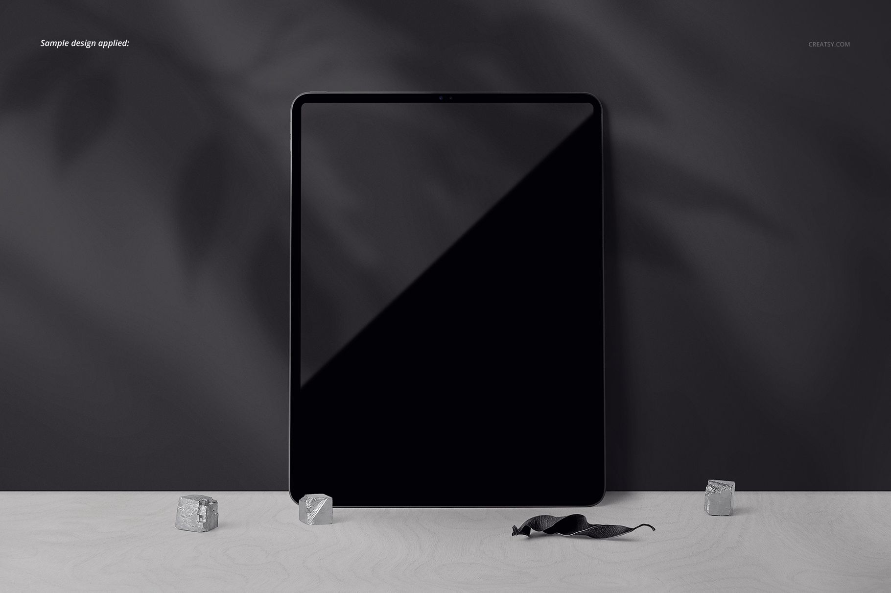 iPad Pro mockup with a blank black display, positioned vertically on a pale wood table, surrounded by stones and a leaf.