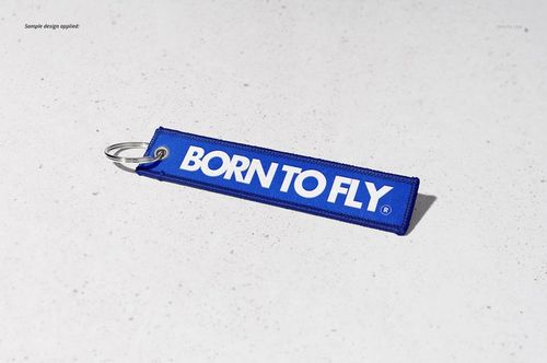 Blue fabric keychain mockup featuring large white lettering, silver ring on one end, displayed on a textured background.