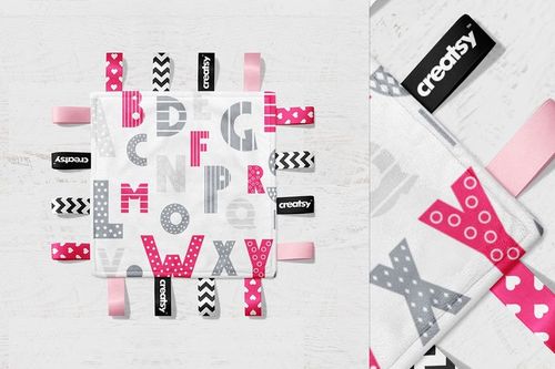 Square taggie blanket mockup with pink and gray alphabet letters, assorted ribbon tags, top view on white wood.