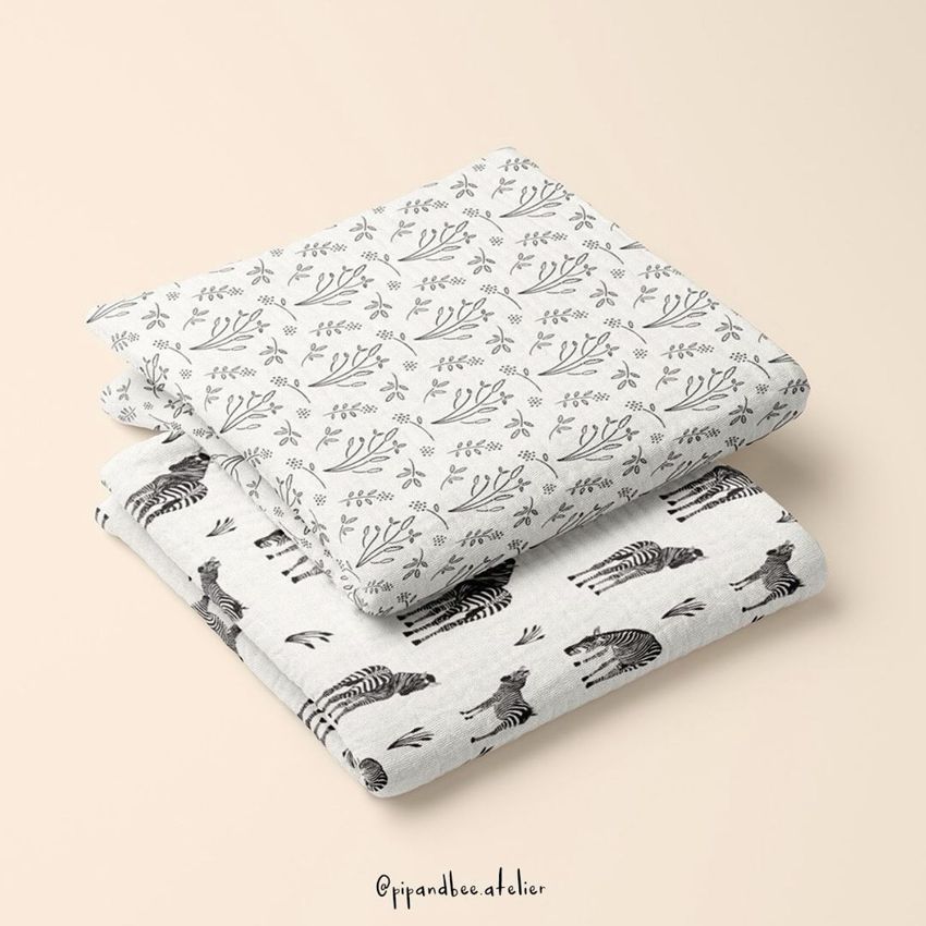 Two crinkle muslin swaddle blankets folded and stacked, featuring black and white botanical and zebra patterns on a beige background.