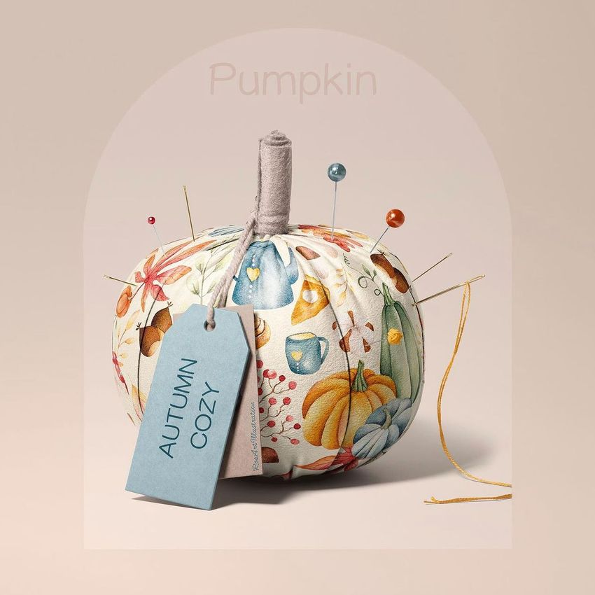 Fabric pumpkin mockup with colorful autumn-themed patterns, sewing pins, and a blue tag reading 'AUTUMN COZY' on a beige background.