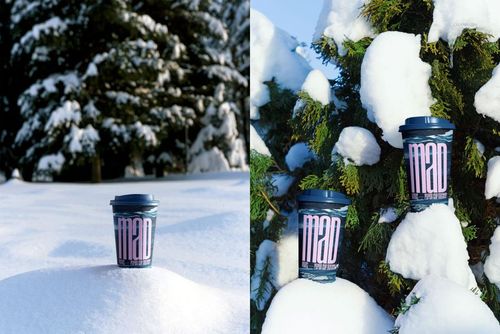 Paper cup mockup with dark lid and bold lettering placed on snowy ground and among snow-covered evergreen branches.