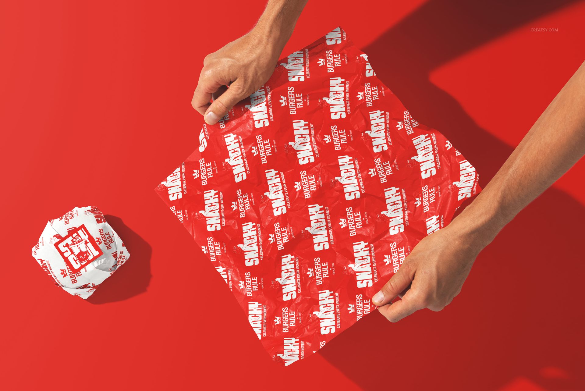 Hands holding a square red burger wrapper with white fast food branding on a vibrant red background, next to a wrapped burger.