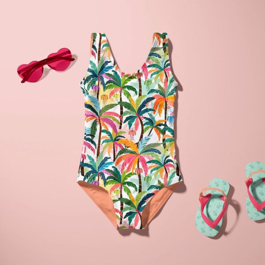 Baby swimsuit mockup with a colorful palm tree pattern, displayed flat on a pink background with sunglasses and flip-flops.
