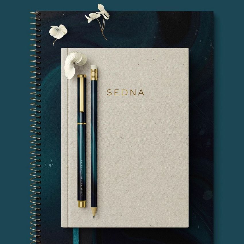 Stationery mockup featuring a beige notebook with gold text, two pens, and white flowers on a spiral-bound pad.