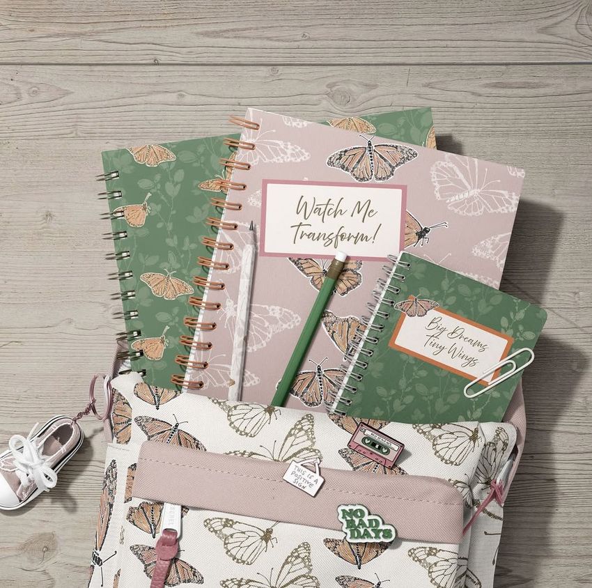 School backpack mockup with spiral notebooks featuring butterfly patterns, arranged on a light wooden surface with a pen and earphones.