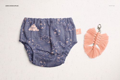 Baby muslin pants mockup featuring a whimsical blue cloud design, shown flat beside a pink tassel accessory.