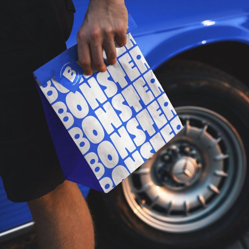 Paper bag mockup with bold blue and white geometric text pattern held by a person standing near a classic blue car wheel.