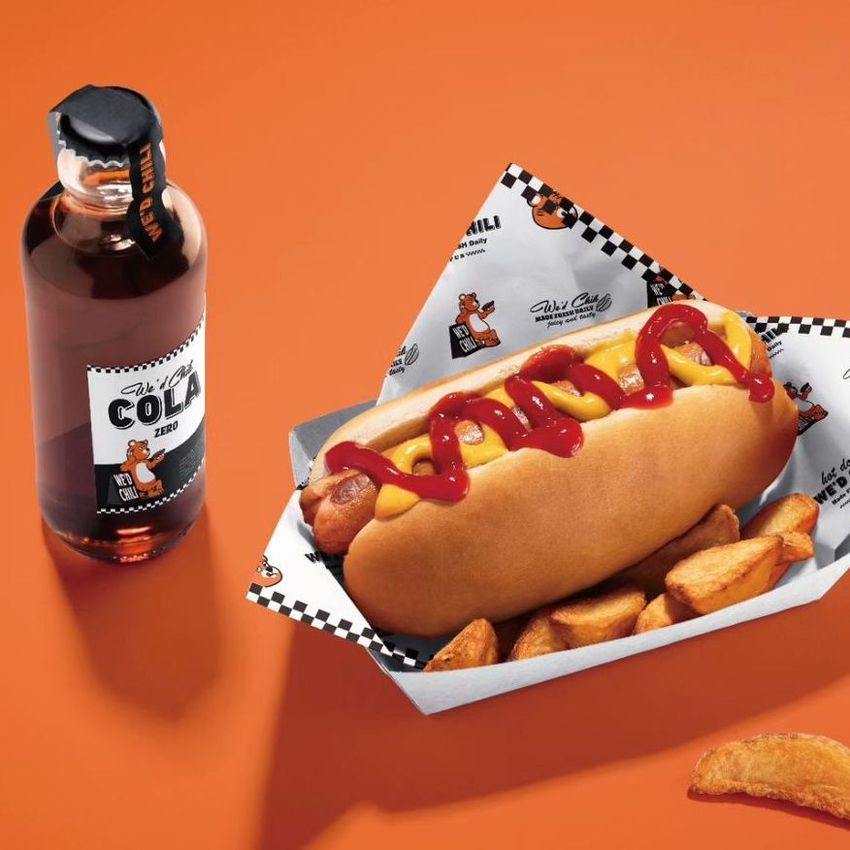 Hot dog mockup wrapped in patterned food safe paper, served with potato wedges on an orange background next to a bottle of cola.