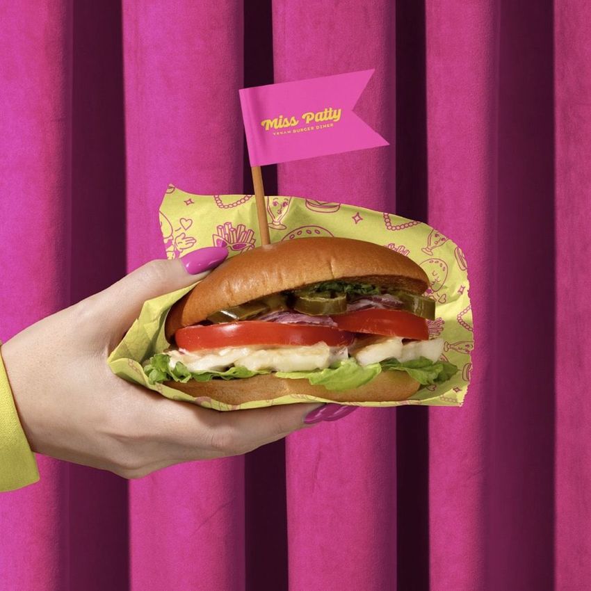 Vegan burger bar packaging mockup featuring a hand with pink nails holding a wrapped burger on a pink curtain backdrop.