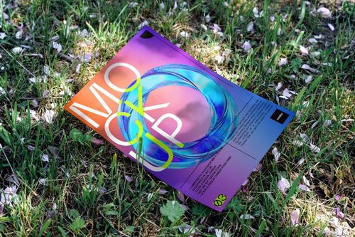 Colorful poster mockup with abstract design lying on grass outdoors, viewed from a slight angle above.