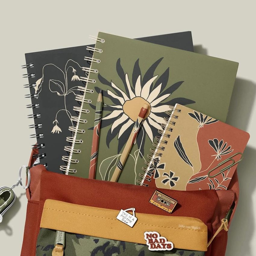School backpack mockup with spiral notebooks featuring wildflower illustrations, pens, and a rust-colored bag on a neutral background.