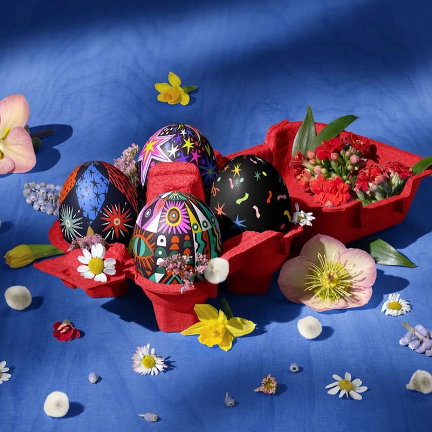 Easter eggs decorated with colorful patterns are arranged in a red carton, surrounded by spring flowers on a blue fabric background.