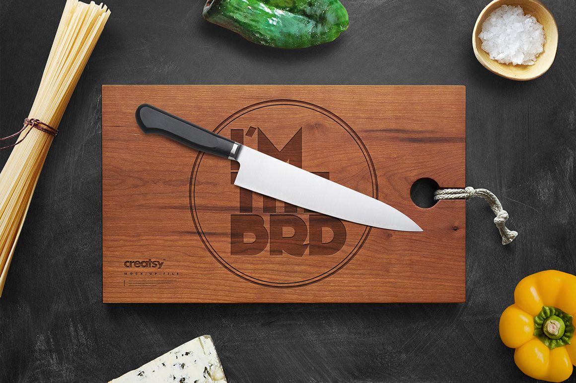Top view of a rectangular engraved wooden board mockup with a chef's knife, surrounded by vegetables and cheese.