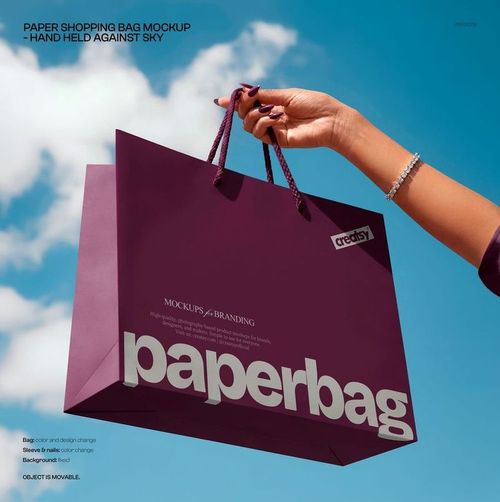 Close-up of a person’s hand grasping a purple paper bag with mockup branding, set outdoors beneath a clear sky.