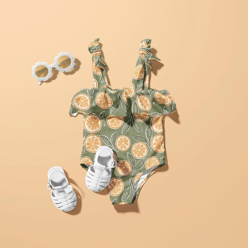 Baby swimsuit mockup with orange slice pattern, styled with white sandals and flower-shaped sunglasses on a peach background.