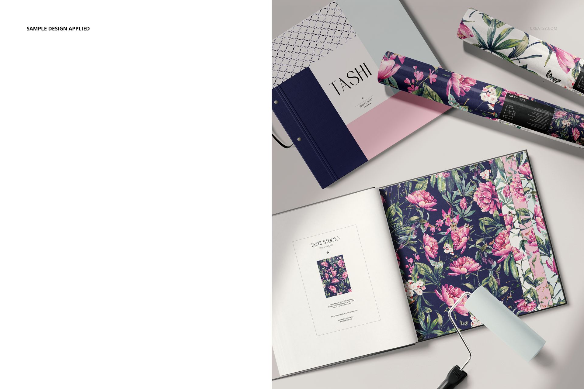 Wallpaper sample book mockup with open pages showing floral patterns, rolled wallpaper, and a paint roller on a light surface.