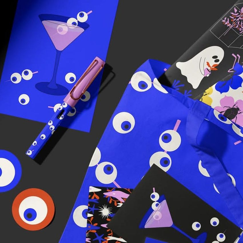Tote bag mockup with bold blue and white eye pattern, surrounded by illustrated stationery items and colorful props on a dark surface.