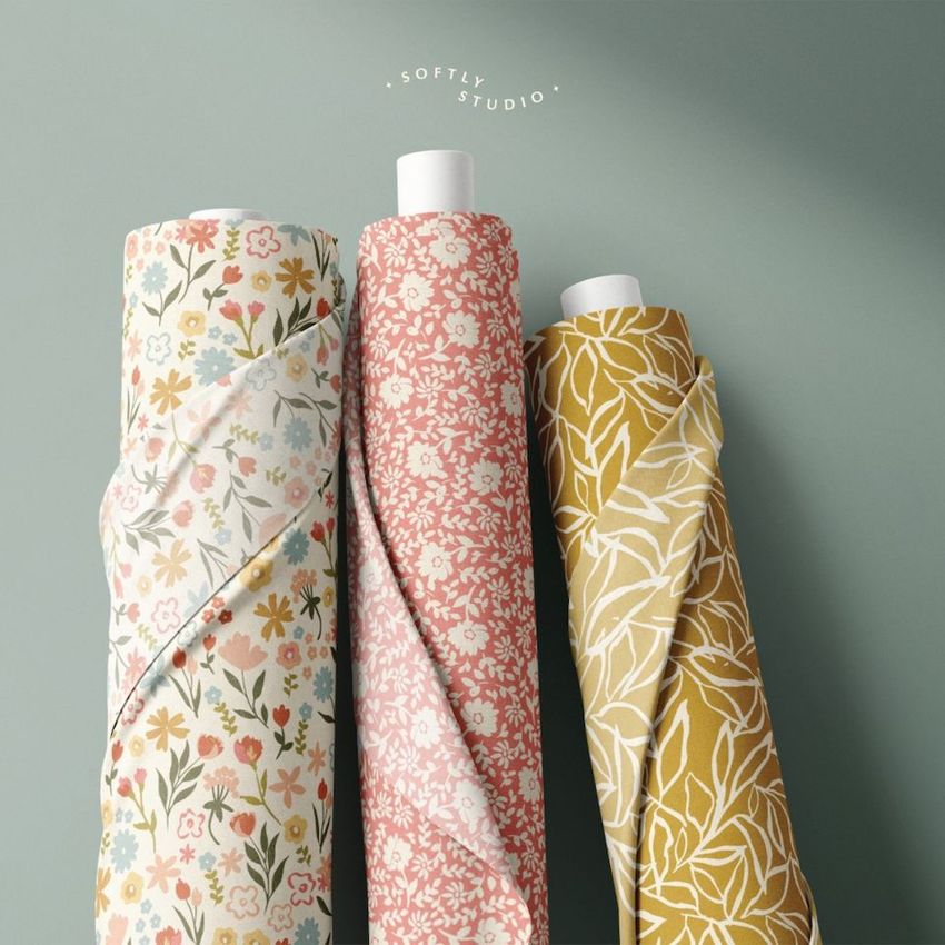 Fabric rolls mockup showing three upright rolls with floral and leaf patterns in pastel, pink, and mustard yellow tones on a plain surface.