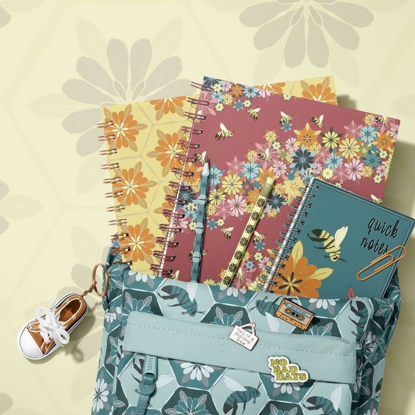 School backpack mockup with a floral pattern, holding spiral notebooks decorated with wildflowers and bees, viewed from above.