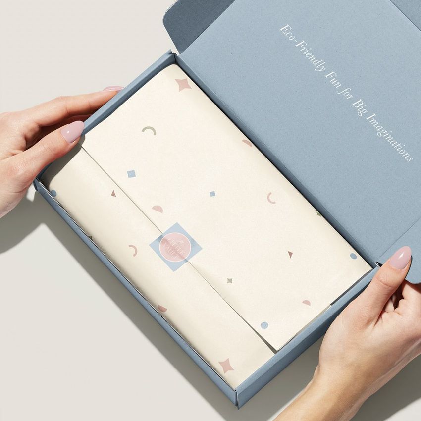 Hands holding open a blue mailing box mockup with patterned tissue paper and a round sticker seal inside, viewed from above.