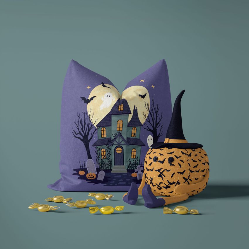 Halloween pillow mockup featuring a haunted house illustration with ghosts and bats, next to a plush toy pumpkin in a witch hat on a plain surface.