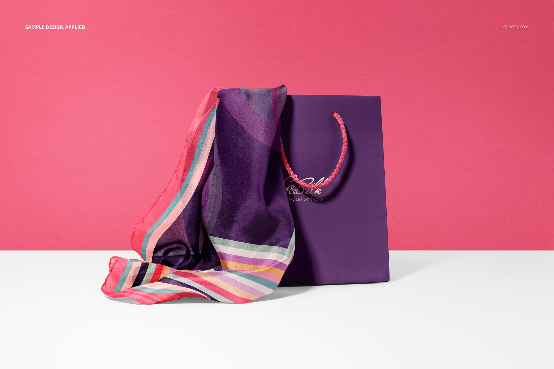 Mockup of a striped silk scarf and purple paper bag arranged on a white surface with a bold pink backdrop.