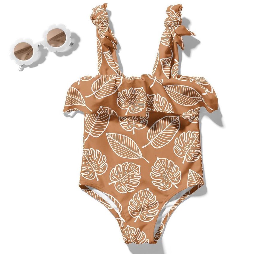 Baby swimsuit mockup featuring a brown one-piece with white tropical leaf pattern, ruffled straps, and matching sunglasses.
