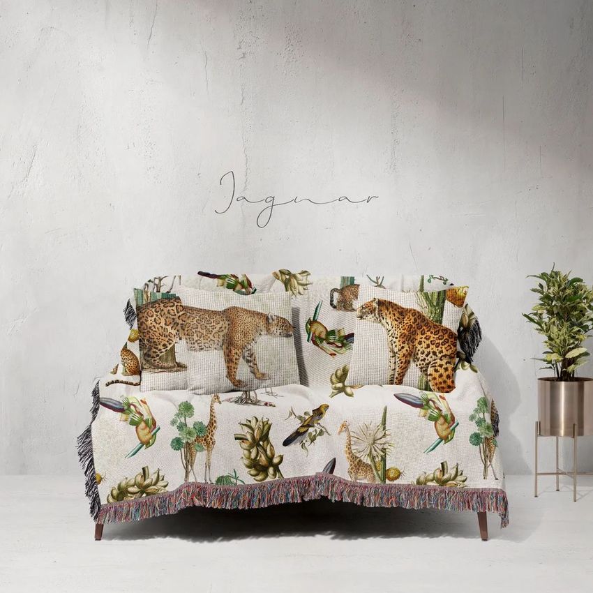 Woven throw blanket mockup with jaguar and tropical plant illustrations draped over a sofa, set against a light textured wall.