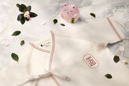 Organic cotton baby kimono bodysuit mockup in cream color with red stitching, displayed on textured fabric with leaves and petals.