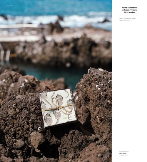 Fabric mockup featuring a square textile piece with a subtle pattern, placed on rugged rocks near blue water.