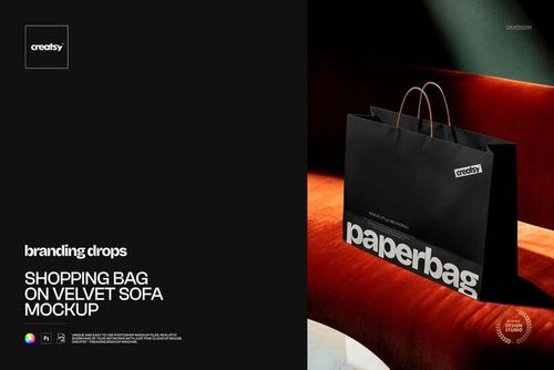 Elegant black paper shopping bag on a deep red velvet sofa, shown in a realistic PSD mockup scene with shadows.