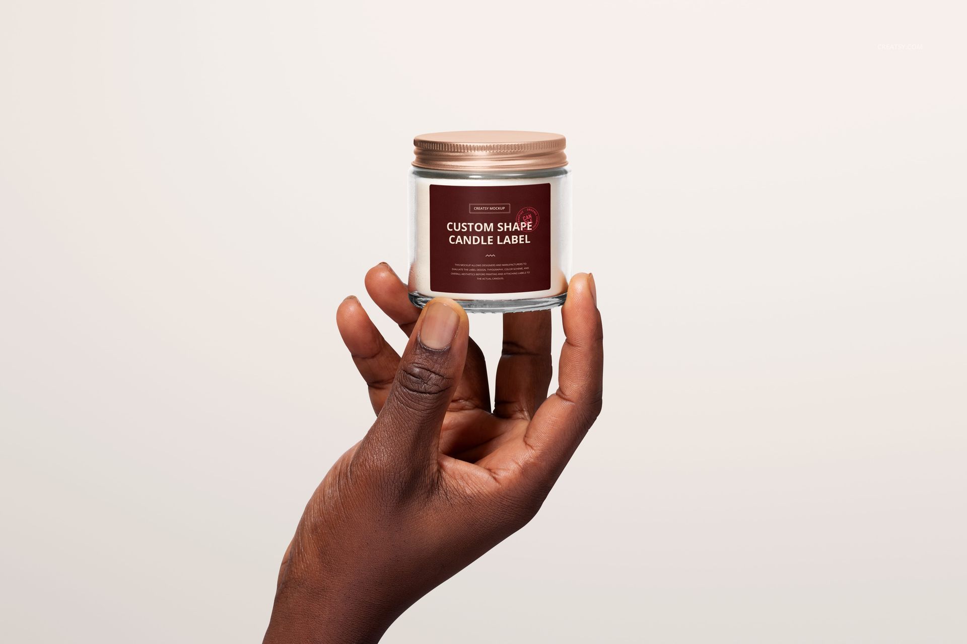 Front perspective of a clear jar candle mockup with custom label, held by hand against a plain off-white background.