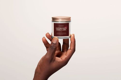 Front perspective of a clear jar candle mockup with custom label, held by hand against a plain off-white background.