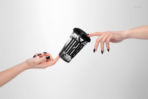 PSD mockup of a paper coffee cup featuring a black lid and abstract pattern, passed between two hands with dark nails.