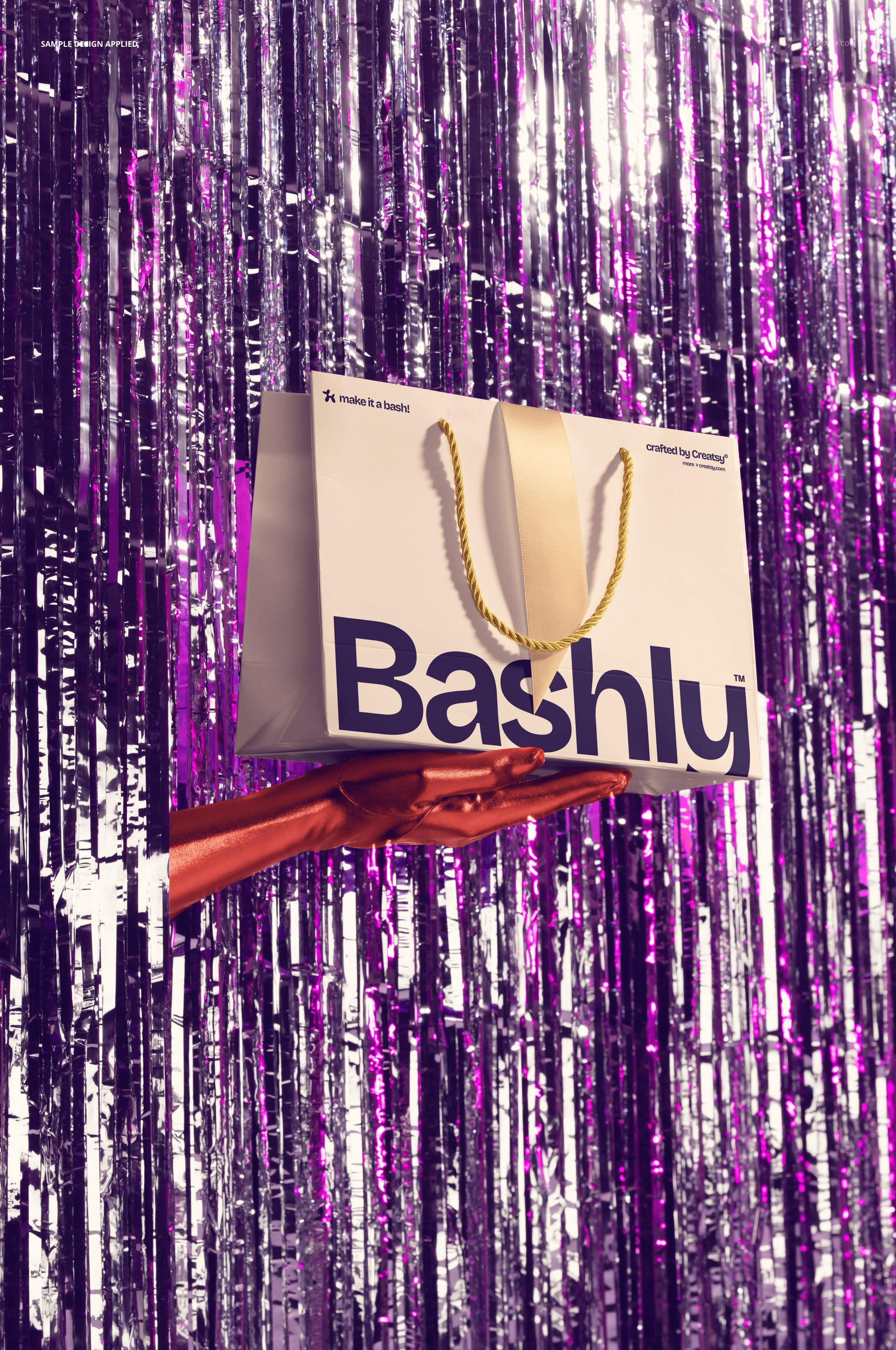 PSD mockup showing a white gift bag with bold text, gold handles, and metallic purple tinsel backdrop, hand holding.