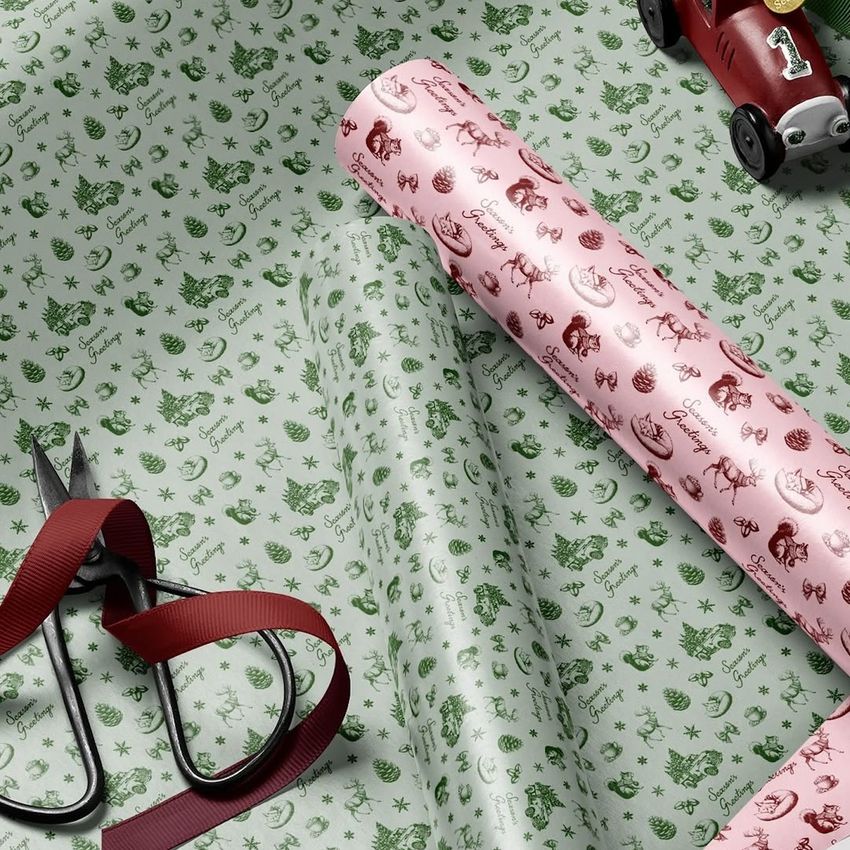 Gift wrapping paper mockup featuring green and pink rolls with Christmas-themed patterns, scissors, and ribbon on a flat surface.