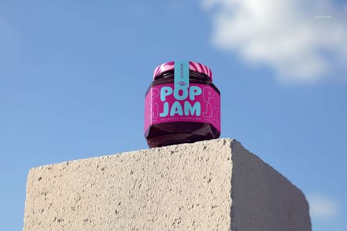 Hexagonal blackcurrant jam jar mockup with bright pink and blue branding, positioned on a light concrete surface against the sky.