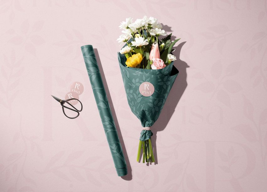 Packaging mockup showing a bouquet of white and yellow flowers wrapped in green floral paper with a round label, next to scissors and a matching paper roll on a pale pink surface.