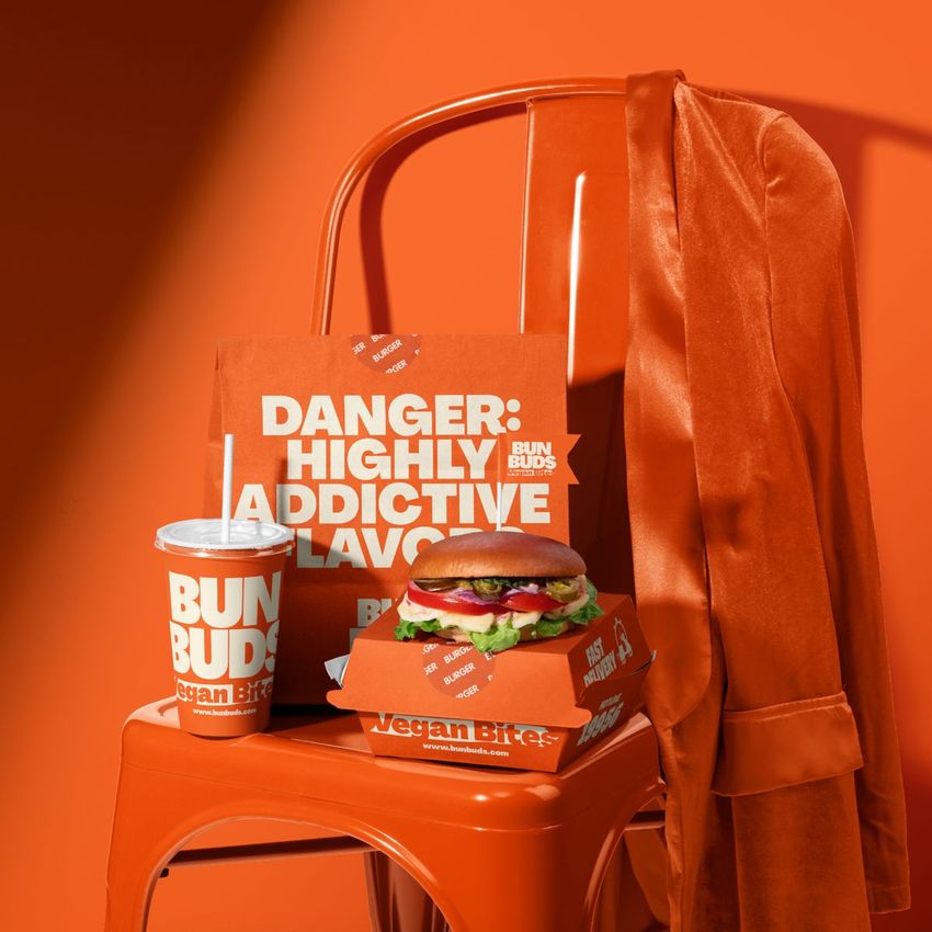 Vegan burger bar packaging mockup featuring a burger in a branded box, paper bag, and drink cup on an orange chair with a jacket.