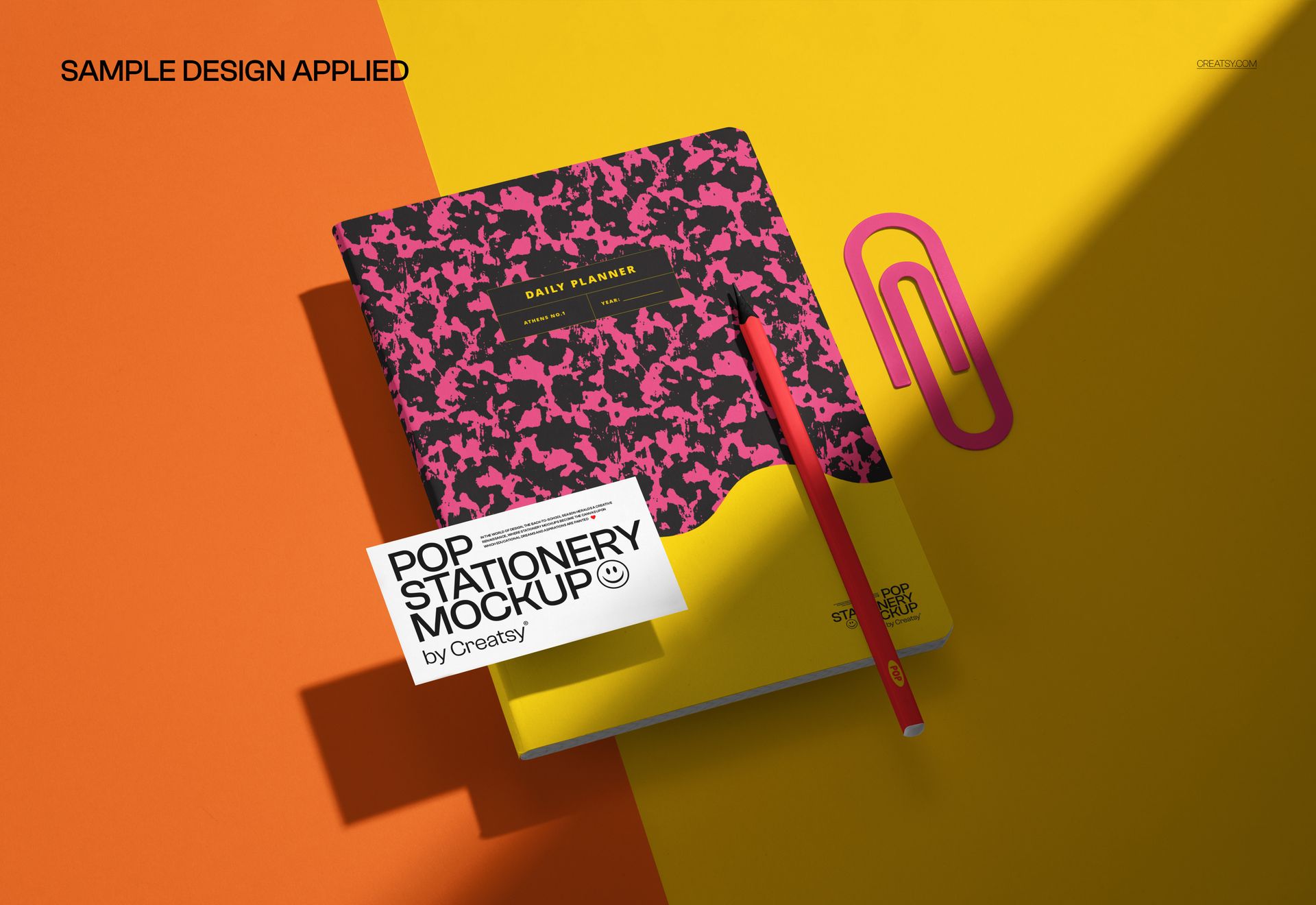 Stationery mockup showing a closed notebook with a colorful patterned cover, a pencil, and a paperclip on a split yellow and orange surface.
