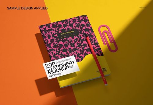 Stationery mockup showing a closed notebook with a colorful patterned cover, a pencil, and a paperclip on a split yellow and orange surface.
