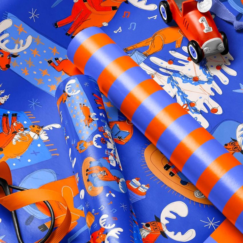 Gift wrapping paper mockup showing close-up rolls and sheets with festive Christmas illustrations in blue and orange tones.