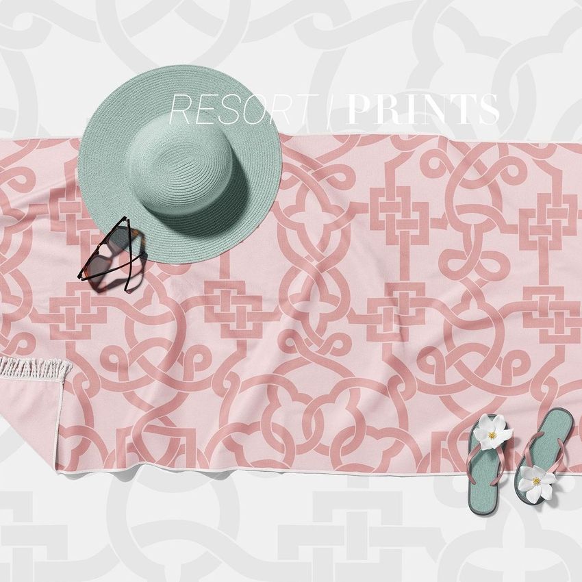 Fringed beach towel mockup with a pink geometric pattern, styled with a wide-brimmed hat, sunglasses, and sandals on a light background.