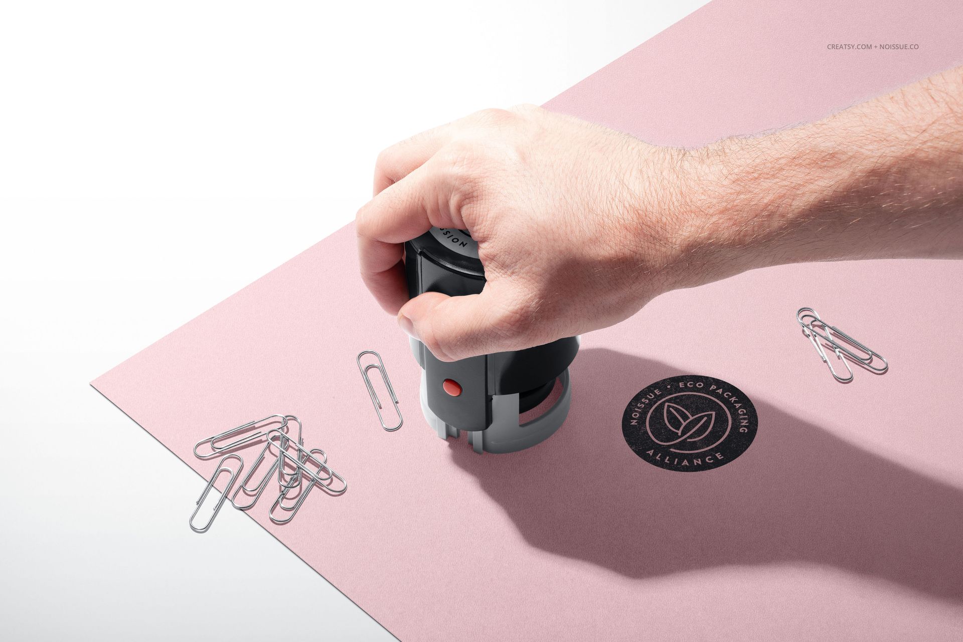 PSD mockup featuring a hand using a black self-inking stamp on pink paper, with scattered paperclips and a visible logo imprint.