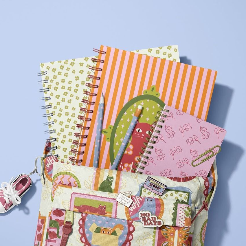 School backpack mockup with spiral notebooks featuring colorful cat illustrations, pens, and a keychain on a light blue background.