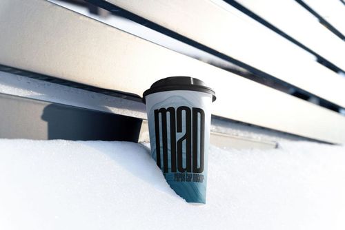 Winter-themed paper cup mockup featuring a black lid and graphic text, placed in snow near a silver bench.