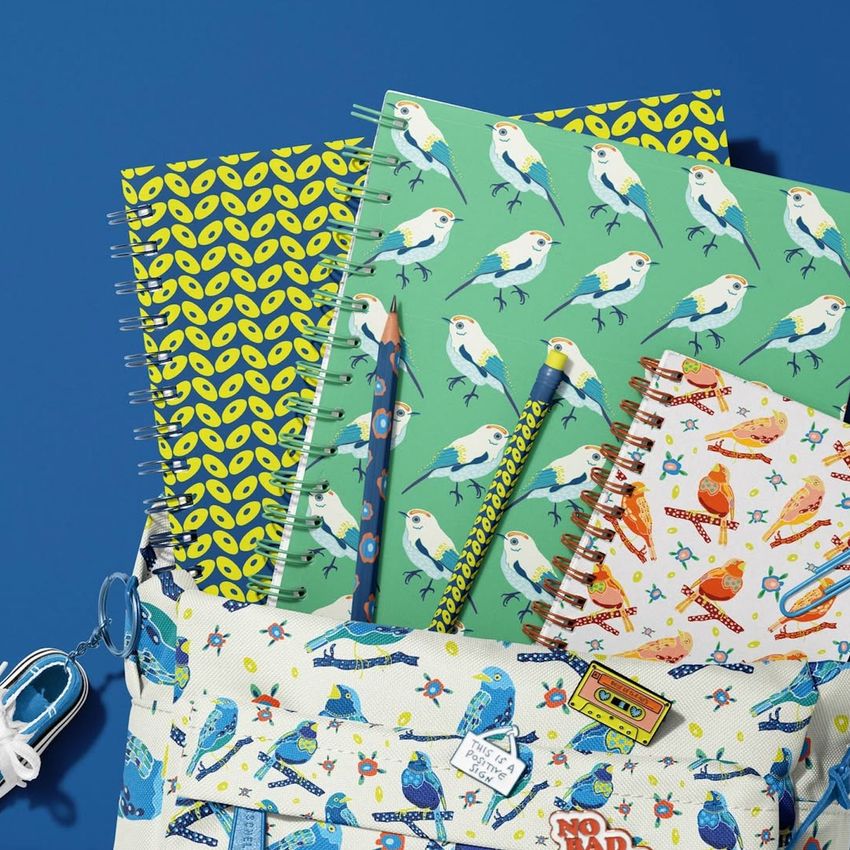 School backpack mockup with spiral notebooks featuring bird and leaf patterns, a pencil, and a keychain on a blue background.